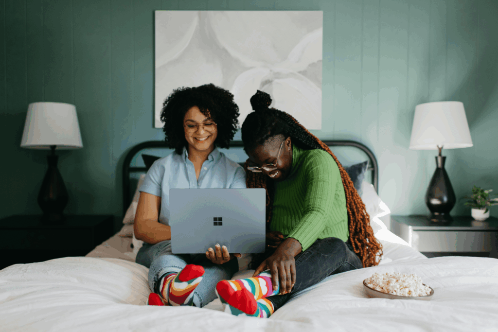 About Us Two women sitting on a bed, smiling while using a laptop together.